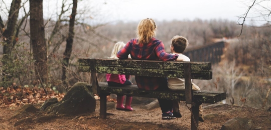 A mother sits on a bench in the woods with her two small children on either side. They are facing away from the camera.